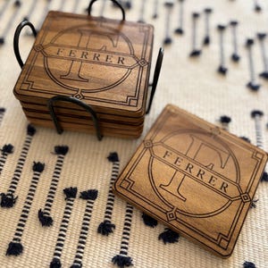 May include: A set of square wooden coasters with a dark brown finish, featuring a laser-engraved design with the letter F and the name FERRER inside a geometric border. The coasters are stacked in a black metal holder with one placed beside it.