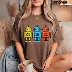 May include: A brown short-sleeve t-shirt featuring a graphic of three cartoon robots in yellow, blue, and orange. Each robot displays binary code on its chest, specifically 01, 10, and 11. The design is centered on the front of the garment.