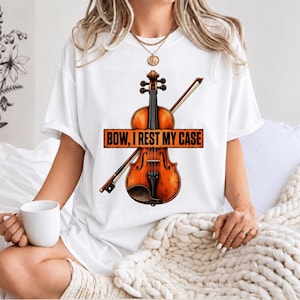 May include: A white short-sleeved t-shirt featuring a graphic of a brown wooden violin and a bow. Across the centre of the violin is an orange rectangular banner with the black text "BOW, I REST MY CASE" printed in a bold, sans-serif font.