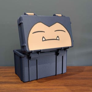 May include: A dark blue rectangular storage case with a hinged lid. The lid features a tan-colored graphic of a sleeping creature face with closed eyes and two small white fangs. The case has a textured matte finish and vertical structural ribs.