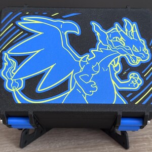 May include: A black rectangular storage case featuring a blue and yellow graphic of a dragon-like creature on the lid. The case has two blue side latches and is supported by a black stand. The design includes diagonal yellow and blue lines.