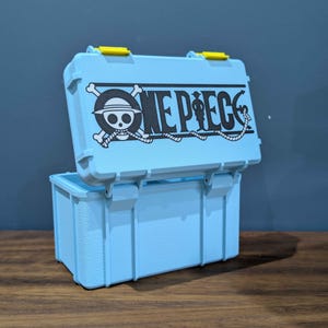 May include: A light blue storage box with a hinged lid and two yellow latches. The lid features a black graphic print of the One Piece logo, which includes a skull and crossbones wearing a straw hat and the text One Piece with an anchor.