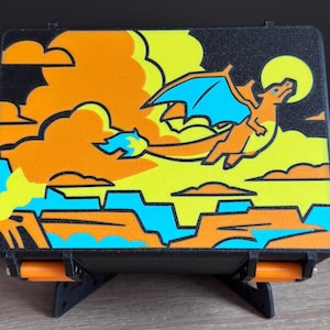 May include: A rectangular black storage box featuring a vibrant graphic print of an orange dragon with blue wings flying against a yellow and orange sky with clouds and a sun. The box has orange hinges and rests on a black stand.