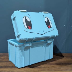 May include: A light blue rectangular storage box with a hinged lid. The lid features a printed graphic of a cartoon face with large eyes and a smiling mouth. The box has a textured exterior surface and two white latches on the top edge.