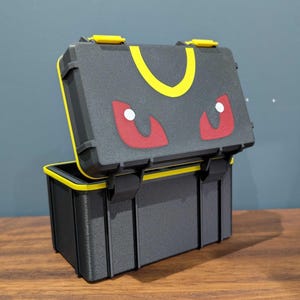 May include: A black rectangular storage case with a hinged lid. The lid features a yellow crescent shape and two red eye-like designs with white pupils. The case has yellow latches and a yellow interior rim, constructed with a textured finish.