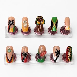May include: A set of ten press-on nails displayed on two clear stands. The designs include palm trees, a dollar sign, a bikini top, the Jamaican flag, leopard print, and a star charm, featuring vibrant colours like green, yellow, red, and black.