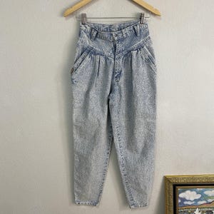 May include: High-waisted acid wash denim jeans featuring a tapered leg silhouette, a button and zipper fly closure, belt loops, and two front pleats. The light blue and white mottled fabric texture is consistent throughout the garment.