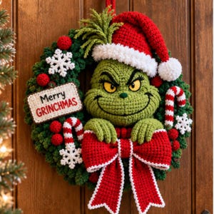 Op de afbeelding: Een ronde groene gehaakte krans met een centraal groen personage dat een rood-witte kerstmuts draagt. De krans is versierd met rood-witte zuurstokken, witte sneeuwvlokken, rode pompons, een grote rode strik en een bordje met de tekst Merry GRINCHMAS.