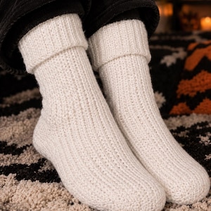 Könnte beinhalten: Ein Paar cremeweiße, wadenlange Socken mit einer dicken, vertikal gerippten Strickstruktur. Die Socken sind mit einem breiten, umgeschlagenen Bündchen am oberen Rand gestaltet, was für einen strukturierten Look sorgt. Das Material wirkt weich.