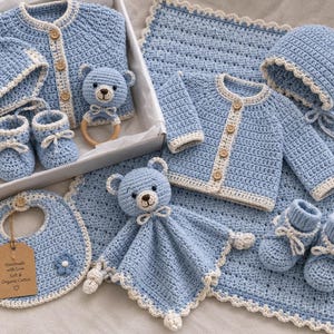 Puede incluir: Un conjunto de regalo para bebé en crochet azul claro que incluye dos cárdigans, dos gorros, dos pares de patucos, un babero, un sonajero de oso y una manta de seguridad de oso. El babero tiene una etiqueta con el texto: Handmade with Love Soft & Organic Cotton.