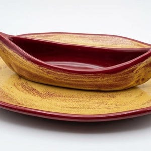 May include: A ceramic serving set consisting of a flat, oval-shaped plate and a matching boat-shaped bowl. Both pieces feature a glossy finish with a yellow base colour and a textured, dark red brushstroke pattern along the edges and interior.