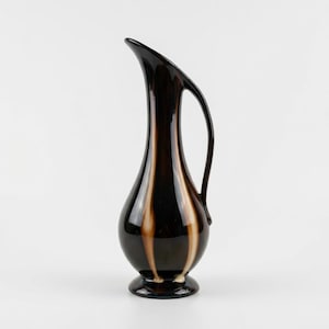 May include: A tall, slender glass pitcher with a dark brown, almost black, glossy finish. The vessel features a bulbous base, a long neck, and a curved handle that extends from the rim to the body, accented by vertical streaks of cream-coloured glass.