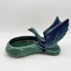 May include: A ceramic swan planter featuring a glossy finish with blended shades of deep navy blue and forest green. The swan is depicted with wings raised in an upward position, attached to an oval-shaped basin that mimics the appearance of water.