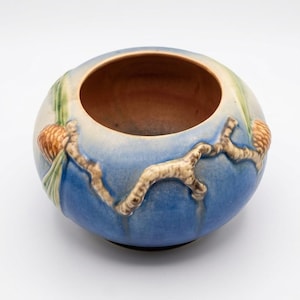 May include: A round ceramic bowl featuring a blue and cream gradient glaze. The exterior is decorated with a raised, textured relief of brown pine branches and orange pinecones with green needles wrapping around the curved surface of the vessel.