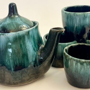 May include: A ceramic tea set consisting of one teapot with a lid and four matching mugs. The pieces feature a glossy glaze with a deep forest green and black drip pattern that transitions into a lighter teal hue toward the top of each item.