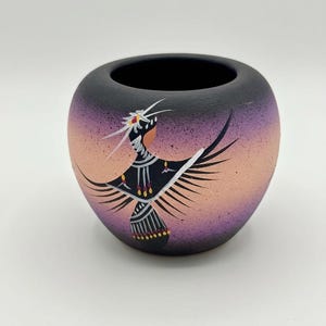 May include: A small, rounded ceramic pot with a matte finish, featuring a gradient colour scheme that transitions from deep black at the top and bottom to a soft peach and purple centre. A stylised black bird figure is painted on the front.