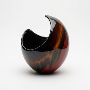 May include: A sculptural ceramic bowl with a smooth, glossy finish. The object features a deep, asymmetrical opening and a rounded base. The exterior displays a blended colour palette of deep black, warm amber, and vibrant red in a drip pattern.