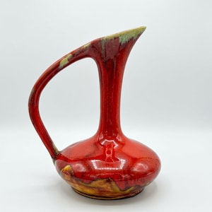 May include: A vibrant red ceramic pitcher with a long, slender neck and a curved handle. The glaze features a glossy finish with subtle yellow and green undertones near the base and rim, creating a smooth, rounded silhouette for this vessel.