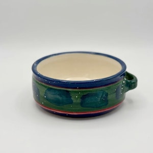 May include: A ceramic bowl with a single handle, featuring a cream-coloured interior and a dark blue rim. The exterior is decorated with a green band, dark teal painted spots, and thin red and blue stripes, accented by small white painted dots.