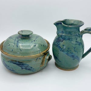 May include: A ceramic lidded bowl and a matching pitcher, both featuring a mottled teal and blue glaze with visible brown clay edges. The bowl has two side handles and a round lid, while the pitcher has a curved handle and a pouring spout.