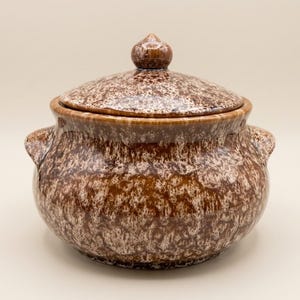 May include: A round ceramic pot with a lid, featuring a glossy brown and white mottled or speckled glaze. The pot has two small side handles and a central knob on the lid. The overall finish is smooth and reflective, showing a consistent pattern.