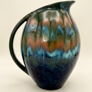 May include: A ceramic pitcher with a glossy finish featuring a drip glaze pattern in shades of deep navy blue, turquoise, and copper brown. The vessel has a rounded body, a curved handle, and a distinct pouring spout at the top rim.
