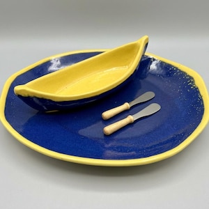 Könnte beinhalten: Ein tiefblauer Keramik-Servierteller mit gelbem Rand, darauf eine passende gelb-blaue, bootsförmige Keramikschale sowie zwei kleine Metallstreicher mit hellen Holzgriffen, die auf dem Teller liegen.