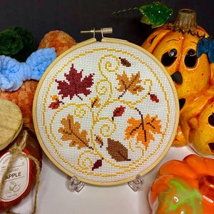May include: A circular wooden embroidery hoop containing a white fabric piece with a cross-stitch pattern of various autumn leaves in shades of red, orange, yellow, and brown, accented with yellow swirling vine designs on a white background.
