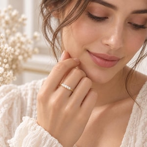 Könnte beinhalten: Ein dünner Goldring mit einer Reihe von fünf kleinen, weißen, ovalen Perlen, die horizontal auf der Ringschiene angeordnet sind. Der Ring wird an einem Finger getragen und zeigt das zarte Metallband sowie den Glanz der schlicht angeordneten Perlen.