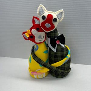 May include: Two stuffed cat figures made of fabric. One cat is yellow with blue and pink floral patterns, wearing a small yellow bow. The other cat is dark green plaid, wearing a black bowtie with a red heart. Both have felt faces.