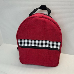 May include: A red backpack featuring a black and white checkered horizontal stripe across the front pocket. The bag includes a black zipper closure on the front compartment and a black fabric handle attached to the top for carrying purposes.