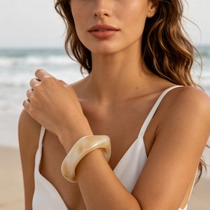 May include: A thick, chunky bangle bracelet with a smooth, polished finish in a marbled cream and beige color. The bracelet features an irregular, organic shape with rounded edges and a glossy, translucent appearance that catches the light.
