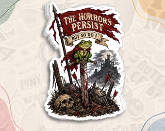 May include: A die-cut sticker featuring a green frog wearing a red cape, perched on a blood-stained sword embedded in the ground. The text reads THE HORRORS PERSIST BUT SO DO I. The scene includes a skull, a helmet, and a dark castle background.