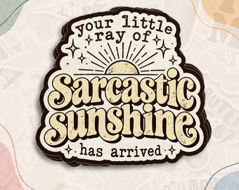 May include: A die-cut sticker featuring a cream-colored background with a sun graphic and the text "your little ray of Sarcastic sunshine has arrived" in dark brown, stylized, retro-inspired lettering with a distressed texture.