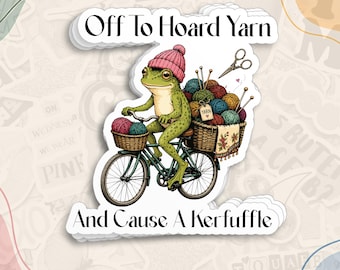 May include: A die-cut sticker featuring a green frog wearing a pink knit beanie, riding a bicycle with baskets filled with colorful yarn balls, knitting needles, and scissors. The text reads Off To Hoard Yarn And Cause A Kerfuffle.