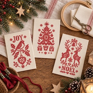 May include: Three rectangular pieces of cream-colored Aida fabric featuring red cross-stitch embroidery. The designs include a bird with the word JOY, a Christmas tree with gifts, and two reindeer with the word NOEL, all in red thread.