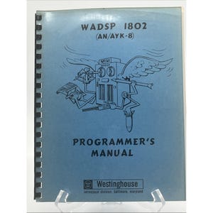 May include: A light blue spiral-bound programmer's manual for the WADSP 1802 (AN/AYK-8) by Westinghouse. The cover features a black line drawing of a winged computer character holding a sextant, a pen, and a bomb, with the title text printed in black.