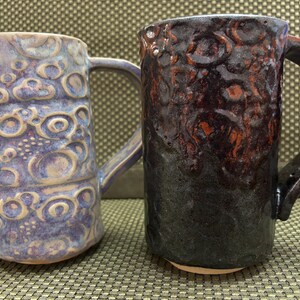 Può includere: Due tazze in ceramica con superfici testurizzate. La tazza a sinistra presenta uno smalto color viola chiaro e crema con motivi circolari in rilievo. La tazza a destra ha uno smalto color marrone scuro e nero con una texture circolare simile.