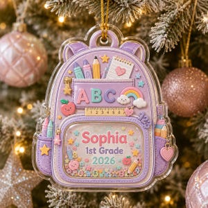 May include: A lavender backpack-shaped Christmas ornament featuring the text 'ABC', '123', 'Sophia', '1st Grade', and '2026'. The design includes a ruler, crayons, a notebook, a rainbow, and small star and heart accents. The center contains loose glitter.