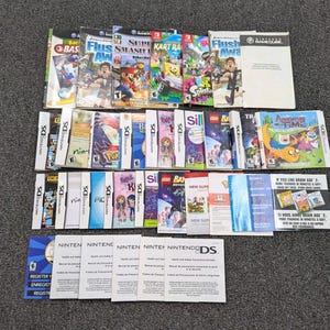 May include: A collection of various video game manuals and inserts for Nintendo GameCube, Nintendo DS, and Nintendo 3DS systems. The assortment includes colorful game booklets, health and safety guides, and registration cards arranged in rows.