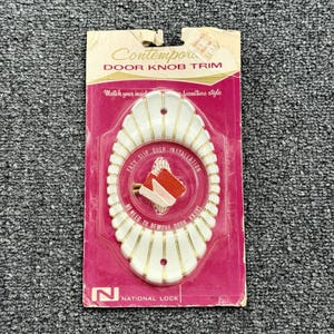 May include: An oval white door knob trim plate with gold-colored vertical stripes, packaged on a pink cardboard backing. The packaging features the text "Contemporary DOOR KNOB TRIM" and "EASY SLIP OVER INSTALLATION NO NEED TO REMOVE DOOR KNOBS."