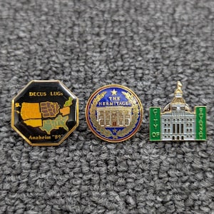 May include: Three metal lapel pins. The first is an octagonal black pin with a map of the United States, gold text reading DECUS LUGS and Anaheim '89. The second is a round blue pin with gold laurel leaves and text reading THE HERMITAGE. The third is a green and white pin shaped like a building with text reading CITY OF SAVANNAH.