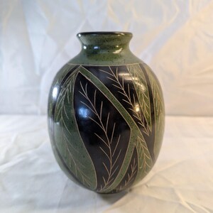 May include: A round ceramic vase with a narrow neck, featuring a glossy finish in shades of deep black and mottled forest green. The surface is decorated with etched, light-colored leaf patterns that contrast against the dark, smooth glaze.