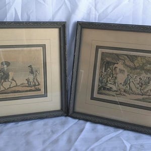 May include: Two rectangular framed prints featuring historical scenes. The left print depicts a figure on horseback near water, while the right print shows a group of people in a landscape. Both are enclosed in dark, textured frames with cream matting.