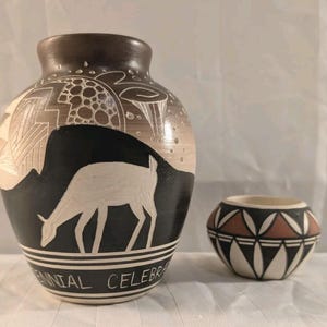 May include: A large, rounded ceramic vase featuring a black and tan gradient with a white etched deer and geometric patterns, alongside a smaller, squat bowl with a black, white, and terracotta geometric design. The vase reads CENTENNIAL CELEBRATION.