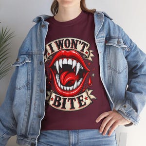 May include: A maroon t-shirt featuring a graphic of red lips with sharp white fangs and a red tongue, surrounded by red blood droplets and two beige banners with the black text I WON'T BITE, worn under a light blue denim jacket.