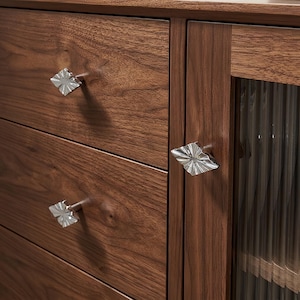 Peut inclure: Un meuble en bois brun foncé doté de deux tiroirs horizontaux et d'une porte latérale avec des panneaux de verre verticaux. Chaque tiroir et la porte sont équipés d'un bouton carré en métal argenté avec un motif texturé en rayons de soleil.