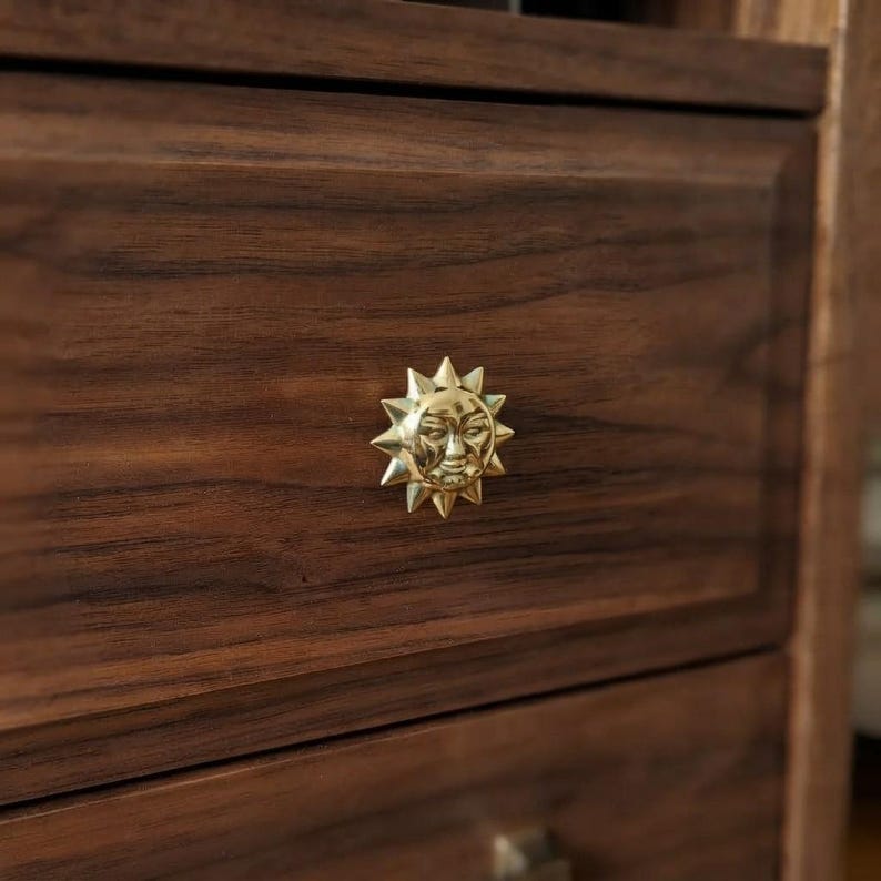 May include: A polished gold-toned metal drawer knob shaped like a sun with a human face, featuring radiating triangular rays. The knob is mounted on a dark brown wooden drawer front with a visible horizontal wood grain pattern and texture.
