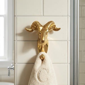 May include: A gold-toned metal wall hook shaped like a ram head with curved horns. The hook features a downward-facing protrusion for hanging items. A beige, textured hand towel with a large embroidered letter C is draped over the hook.