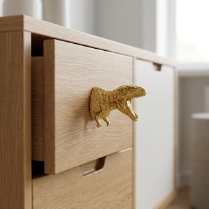 May include: A gold-coloured dinosaur-shaped drawer pull attached to a light oak wood drawer. The dinosaur figure is depicted in a roaring position with its mouth open, featuring textured skin details and small front limbs extending from the drawer face.
